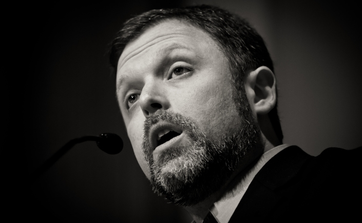 A black and white photo of Tim Wise speaking into a microphone in front of a dark background.
